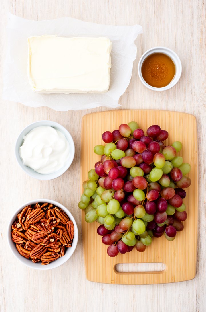 Overhead flat-lay of grape salad ingredients arranged on a light wood surface — a block of cream cheese on parchment, a small bowl of sour cream, a ramekin of vanilla extract, a pile of red and green 