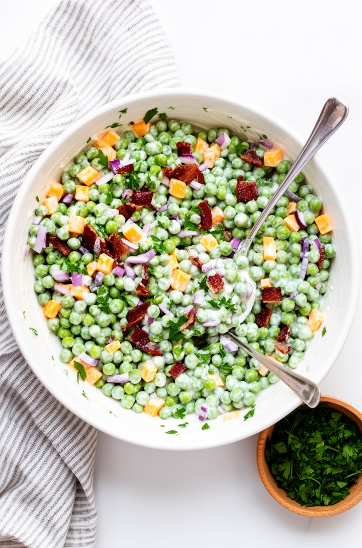 Overhead shot looking directly into a large white ceramic bowl filled with freshly assembled pea salad — bright green peas coated in creamy white dressing, visible orange cheddar cubes and dark crispy