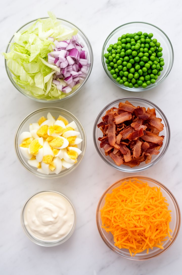 Flat-lay overhead shot of all seven layer salad ingredients arranged in separate small glass bowls on a white marble countertop: a bowl of shredded iceberg lettuce, diced red onion, bright green thawe