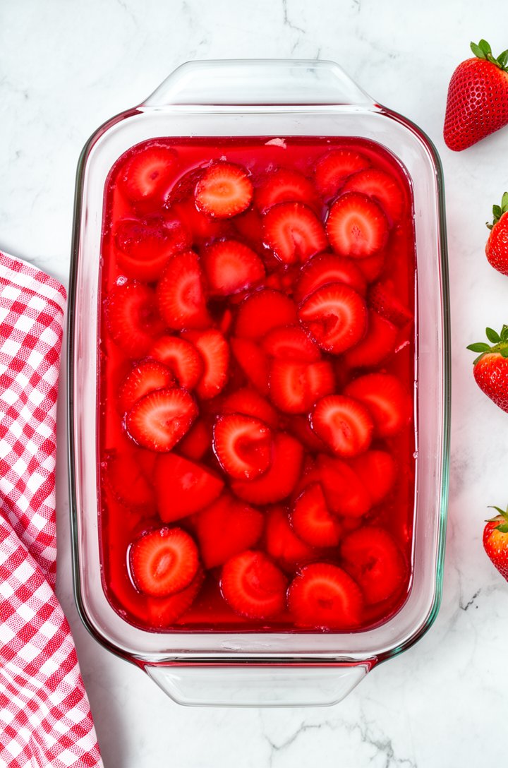 Overhead shot looking down into a 9x13 glass baking dish filled with set strawberry pretzel salad, the surface a glistening ruby-red jello with sliced strawberries visible throughout arranged in overl