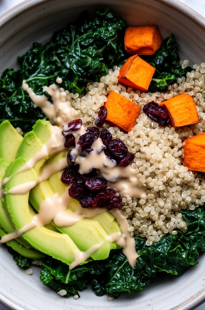 Extreme close-up overhead of a grain bowl style salad with dark green massaged kale, white quinoa, orange roasted sweet potato cubes, sliced avocado showing creamy green interior, dried cranberries, d